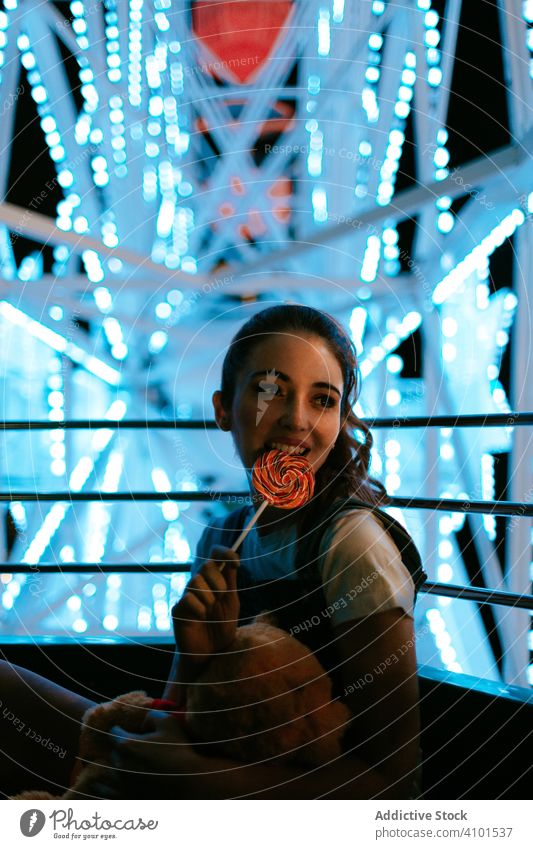 Millennial woman sitting in Ferris wheel and eating lollipop carousel ferris wheel amusement park sweet cheerful night teenager millennial candy toy female