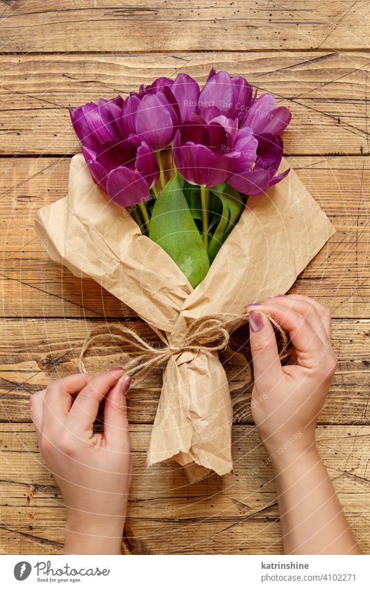 Hands with purple tulips bouquet in craft paper on wooden table hand flowers top view hold spring wedding bow prepare create valentine present violet kraft