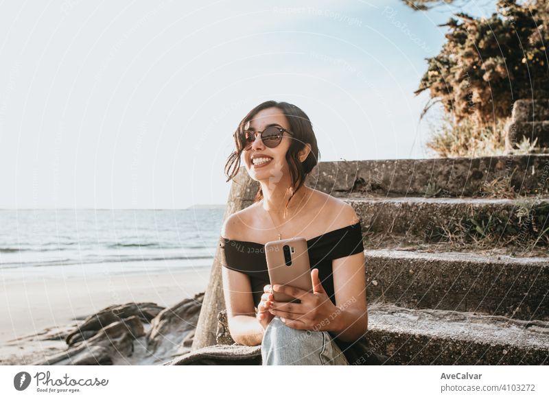 Young woman on summer clothes and sunglasses with his phone smiling with copy space at the beach female african afro american smile person face happy young