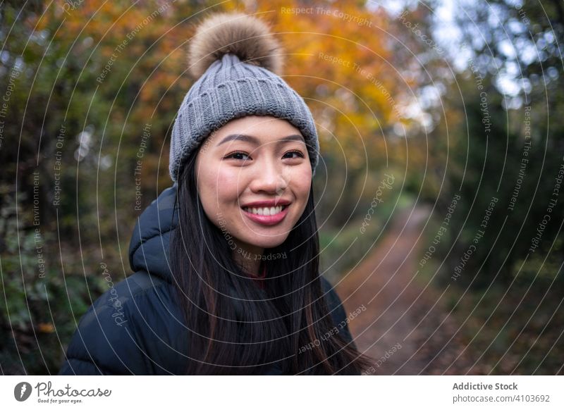 Smiling Asian woman in warm clothes standing in park with trees and stony fence garden stylish autumn nature road path leaves street hat san marino italy pompom