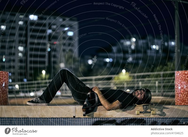 Male skateboarder lying on fence in city at night man rest relax helmet street evening young modern contemporary active male urban illumination building sport
