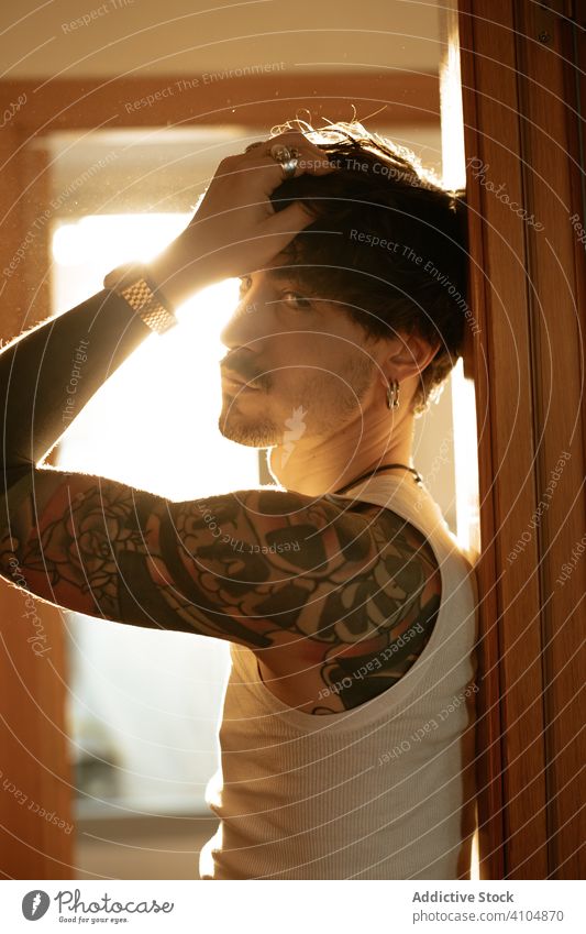 Male leaning on wall and looking at camera man informal tattoo cool brunet mustache handsome ring peek tank top male stylish trendy earring soulful shadow shade