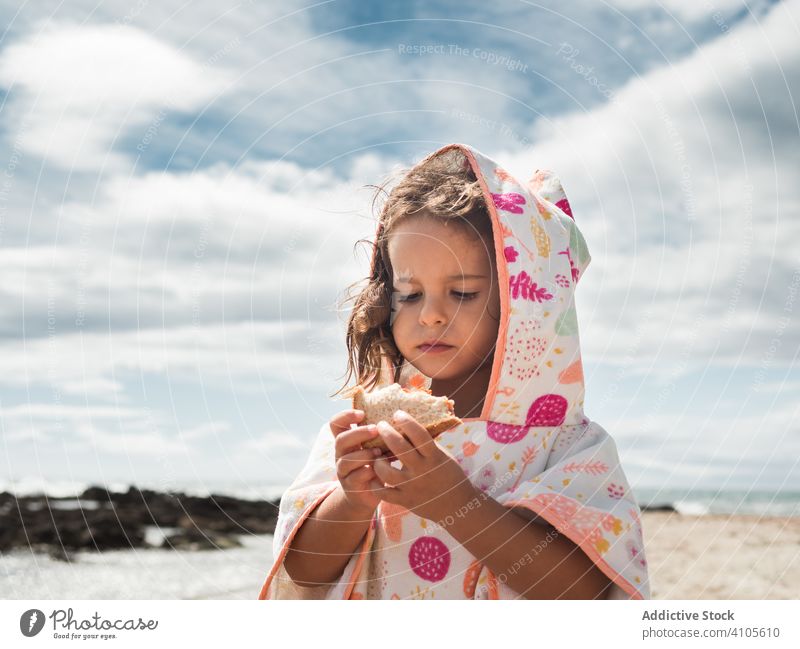 Little girl eating bread on beach summer sunny daytime cloudy sky kid child hood apparel garment pastry bun piece fresh chew bite food lifestyle rest relax