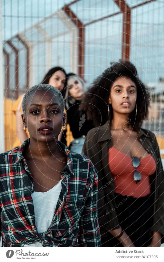 Ethnic young women in casual clothes looking at camera with fearless eyes female friend hipster group serious urban standing diverse style fence multiracial