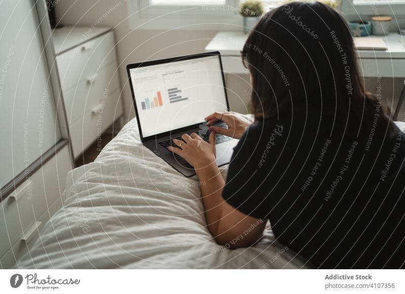 Focused young female student using laptop on bed at home woman typing study lying education communication connection contact internet online surfing browsing
