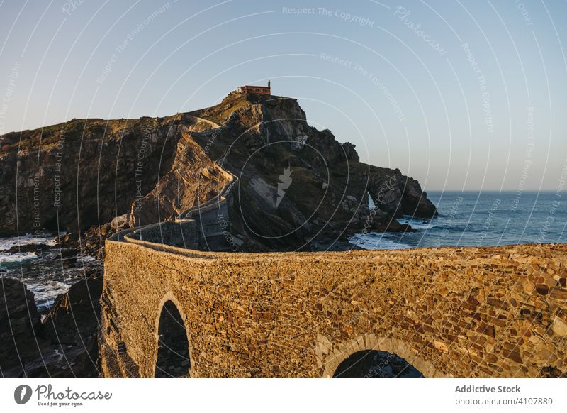 Road going along stone bridge and rocky hill to solitary building on cliff against calm ocean water during sunrise road sea house island spain gaztelugatxe wave
