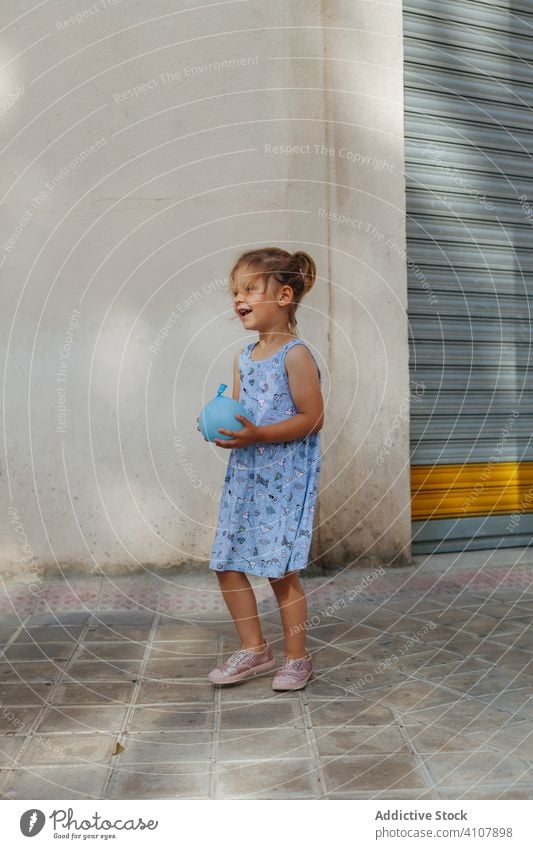 Girl with water balloon on street girl sidewalk summer water bomb play town sunny daytime kid child city pavement toy cute adorable childhood innocence weekend