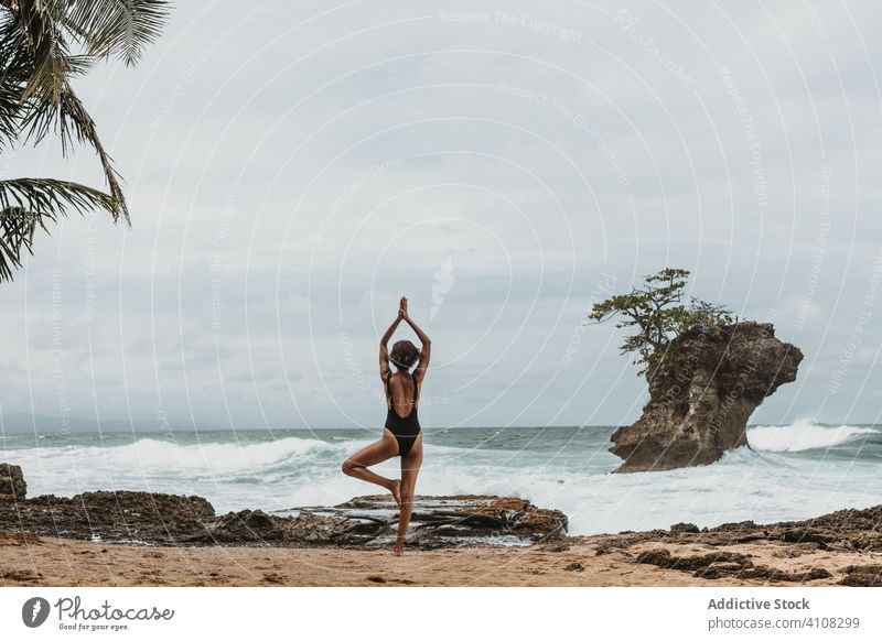 Anonymous lady in swimwear doing yoga on stormy seashore woman meditate ocean balance harmony power beach arm raised swimsuit freedom summer sky resort female