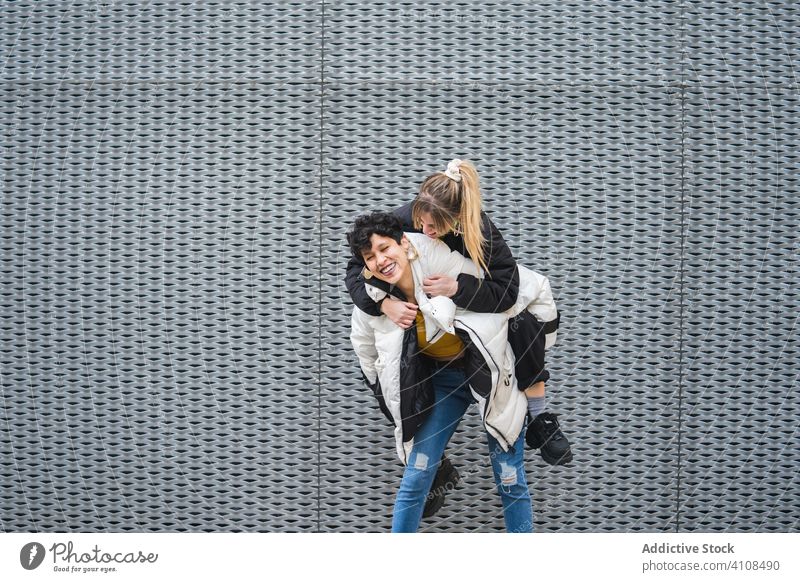 Contemporary female teenagers having fun together women friend piggyback happy laugh urban trendy hipster smile enjoy city street wall building casual stylish