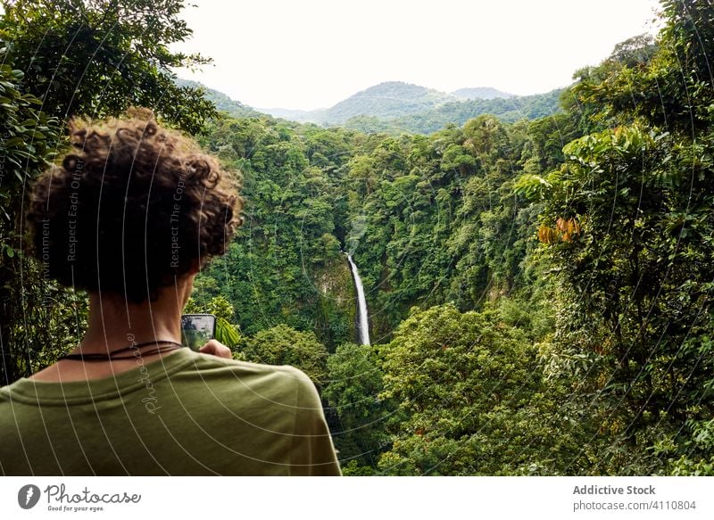 Anonymous traveler using smartphone near waterfall man jungle lush green nature costa rica male trip journey explore visit device gadget browsing cascade online