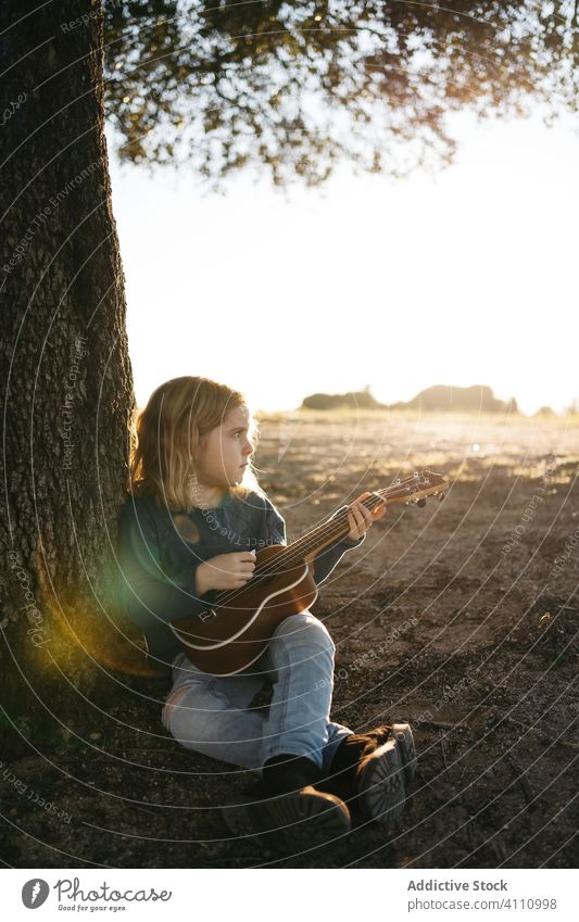 Little girl sitting under tree and playing ukulele kid music guitar nature child summer serious little instrument sound song melody trunk perform practice rest