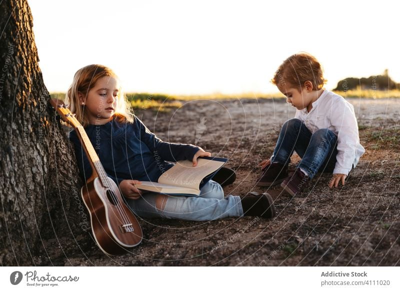 Kids sitting under tree and reading book children nature together summer kid ukulele guitar rest tranquil sister brother sibling friend countryside idyllic
