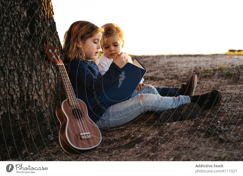 Kids sitting under tree and reading book children nature together summer kid ukulele guitar rest tranquil sister brother sibling friend countryside idyllic