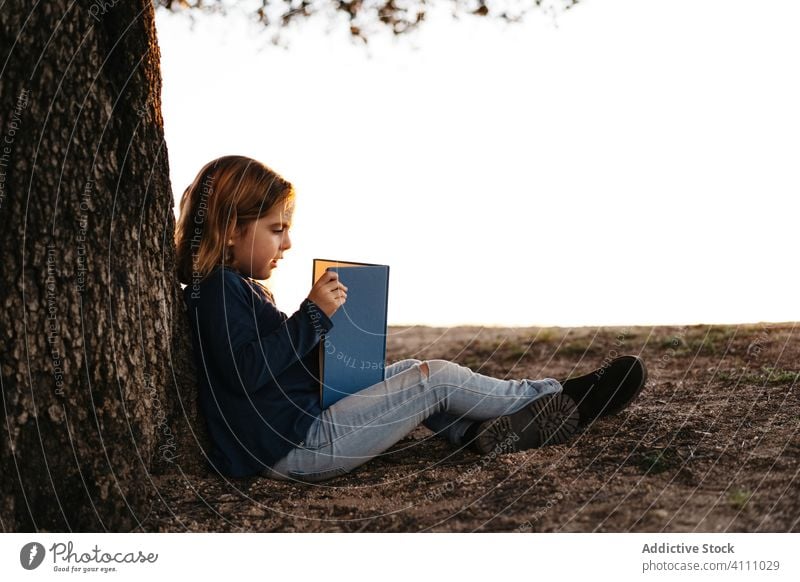 Little girl with book sitting under tree kid read nature field trunk summer casual child calm tranquil serious concentrate female learn meadow lifestyle