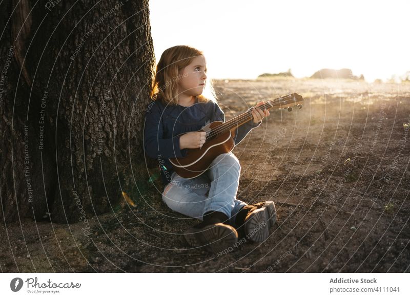 Little girl sitting under tree and playing ukulele kid music guitar nature child summer serious little instrument sound song melody trunk perform practice rest