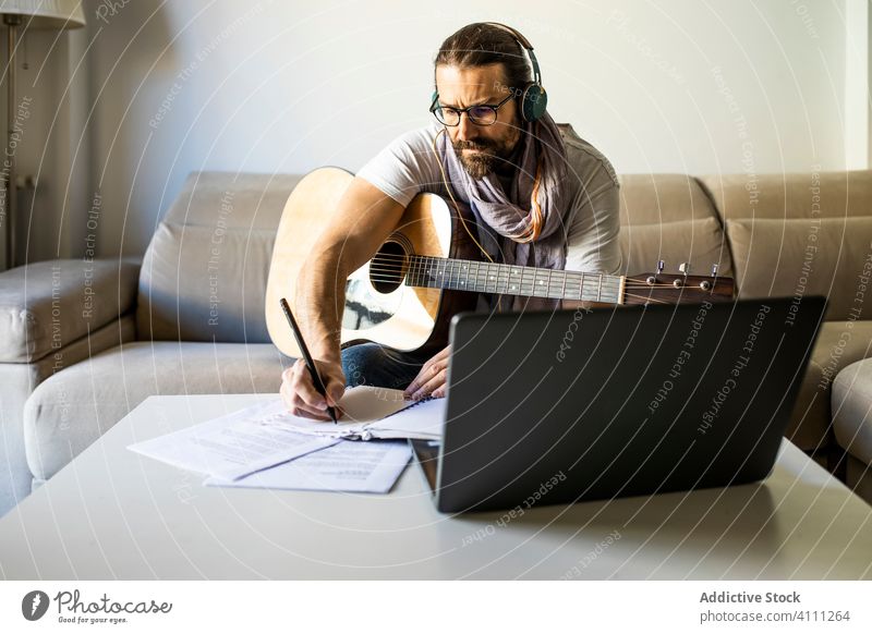 Contemporary man with guitar writing chords in living room musician laptop headphones couch write note songwriter composer poet male using beard contemporary
