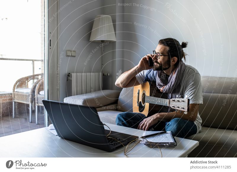 Young male musician calling on smartphone in living room talk guitar using browsing laptop sit couch songwriter composer poet sofa casual communicate man hobby