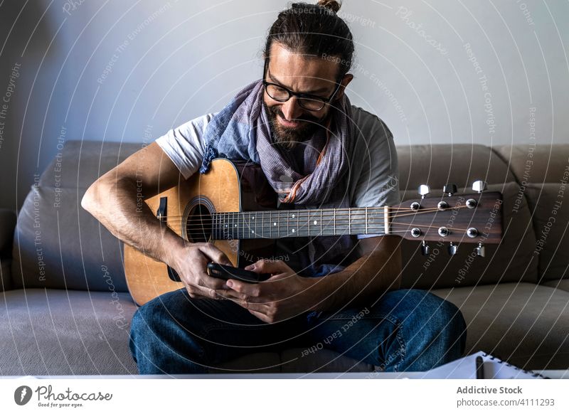 Cheerful man with guitar checking smartphone on sofa sit couch using browsing smile male casual musician mobile beard laugh enjoy watch surfing connection