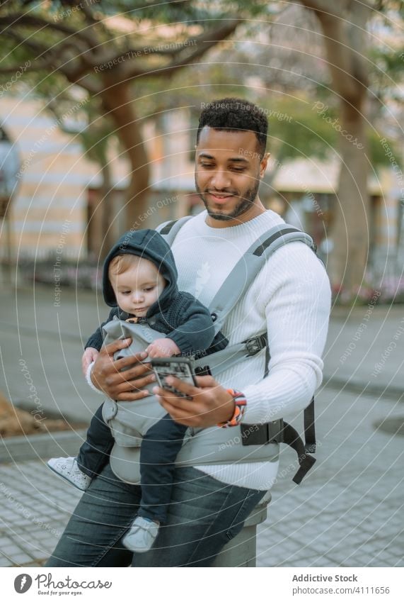 Happy black man with baby in carrier using phone on street father smartphone texting urban modern dad child infant happy son sling device smile parent surfing
