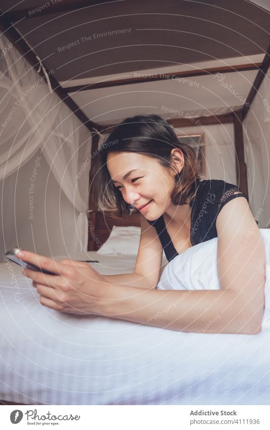 Happy Asian woman using smartphone in bed happy smile ethnic pillow home cozy female relax rest bedroom young comfort device gadget browsing watch lifestyle