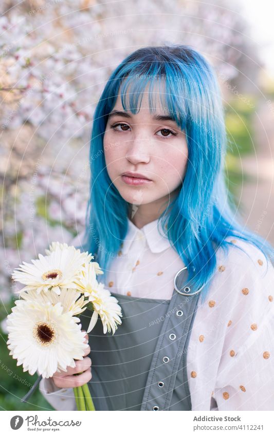 Stylish young woman with flowers standing in blooming garden spring melancholy informal alternative style trendy blue hair modern dyed hair emotionless blossom