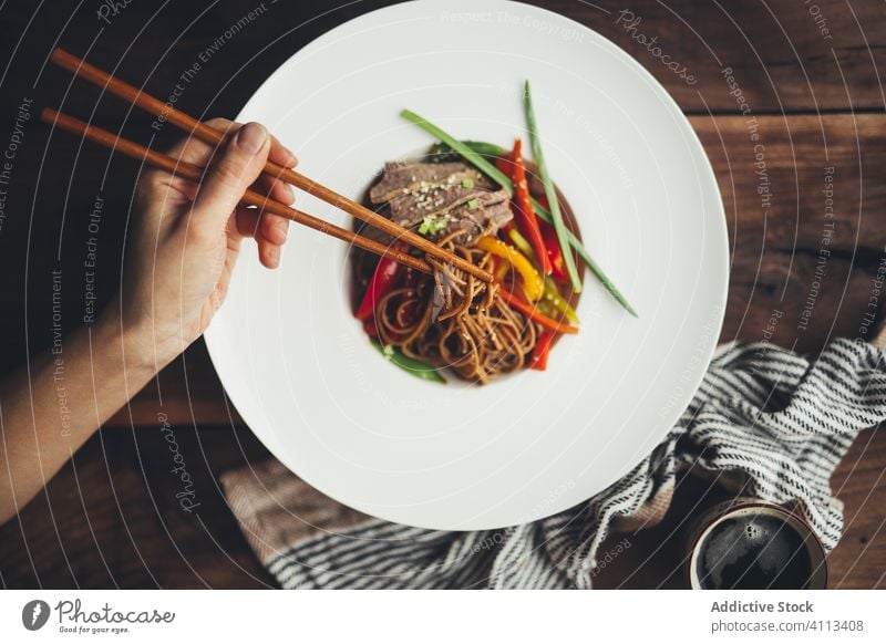Person eating Asian noodles with chopsticks food dish asian hand meat vegetable tradition meal delicious wooden white plate dinner tasty lunch cuisine healthy