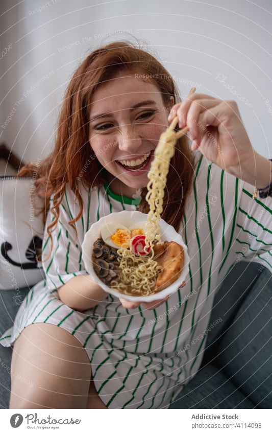 Happy woman eating noodles at home ramen happy japanese sofa chopstick young female redhead cheerful food tradition authentic asian oriental laugh couch tasty