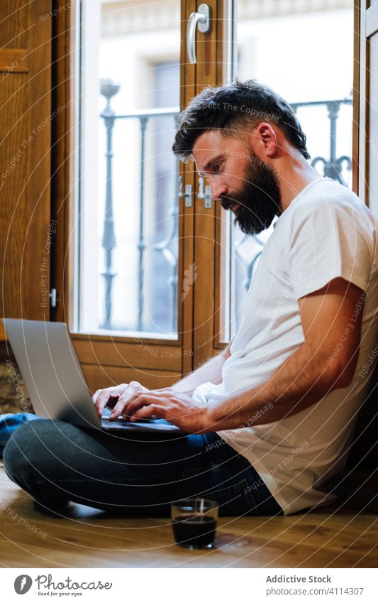 Bearded freelancer using laptop at home man typing drink floor project modern male casual device work internet lifestyle gadget browsing data guy startup remote