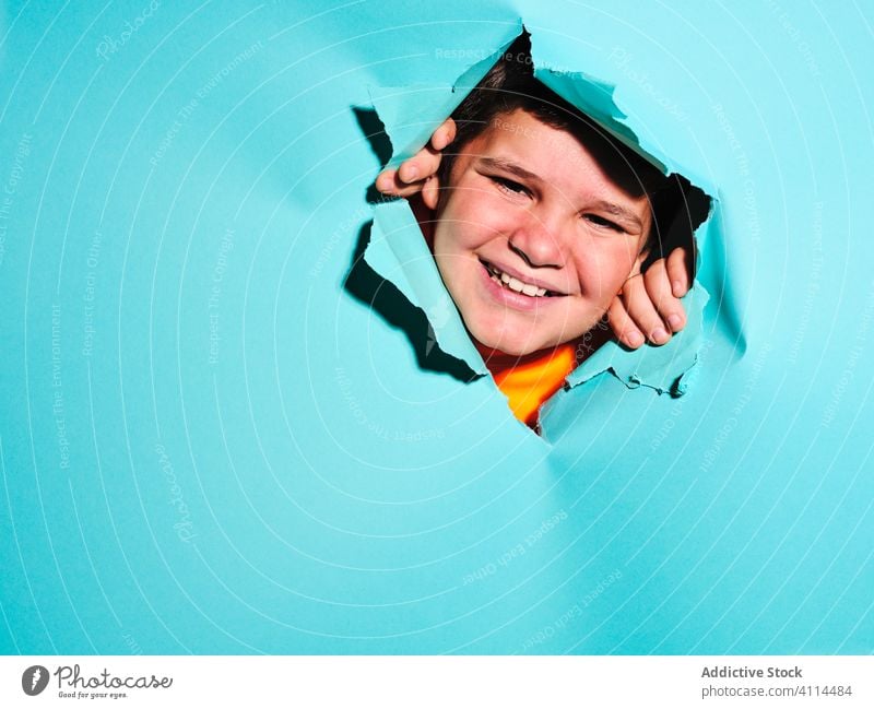 Happy boy looking through paper hole kid grimace ripped expressive concept child male copy space looking at camera blue background personality plus size