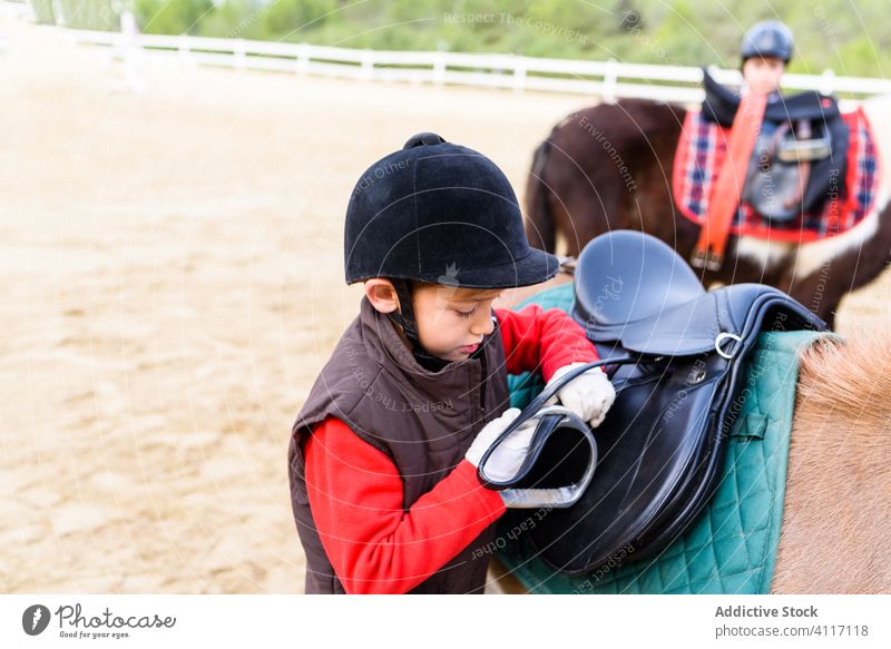 Boy adjusting stirrup on saddle boy pony equestrian school arena dressage jockey kid child lesson horseback paddock fasten helmet little animal ride prepare