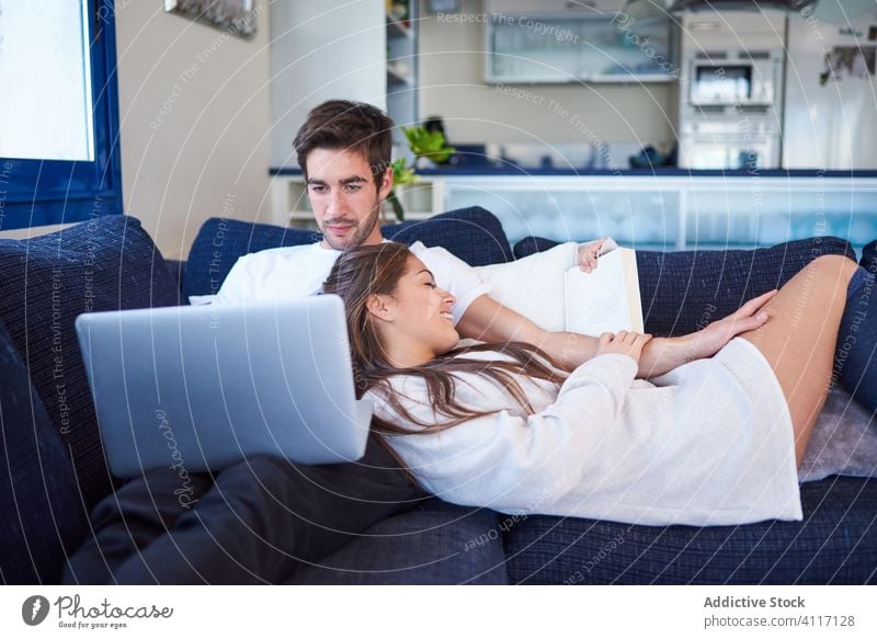 Young couple resting on cozy sofa at home together book laptop using read young relationship husband couch device gadget online internet calm tranquil fondness