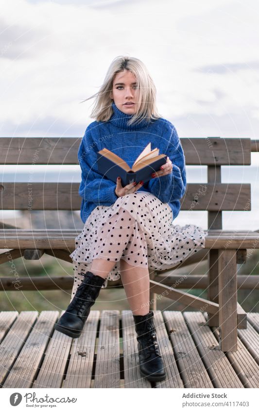 Young woman reading book on seashore terrace bench beach rest young blue sweater nature relax female wooden style vacation enjoy holiday modern trendy blond