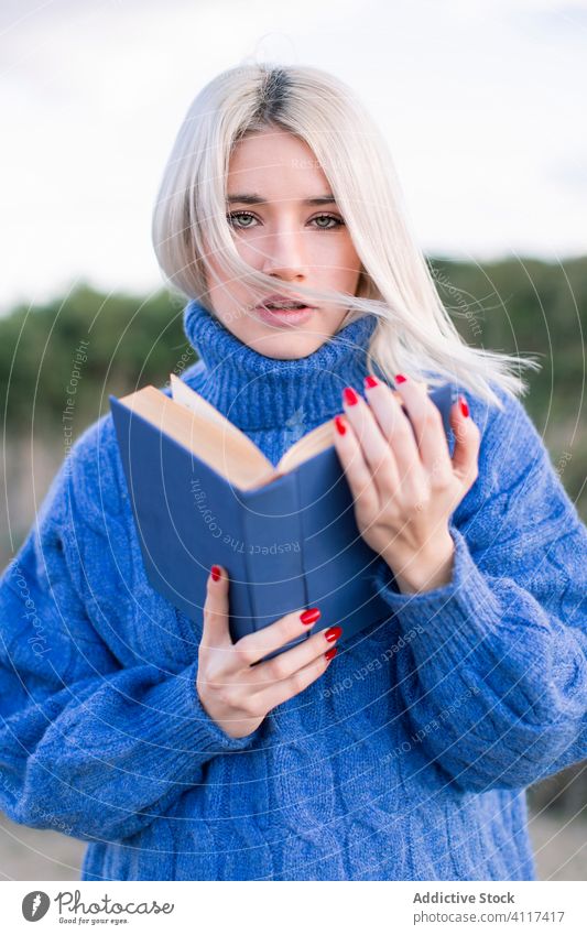 Young woman with book looking at camera blue sweater read young casual serious nature blonde confident modern female education hobby student rest smart learn
