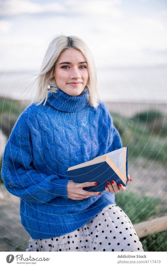 Woman enjoying book in countryside looking at camera woman read happy cheerful nature sweater young female casual blond rest literature hobby smile charming