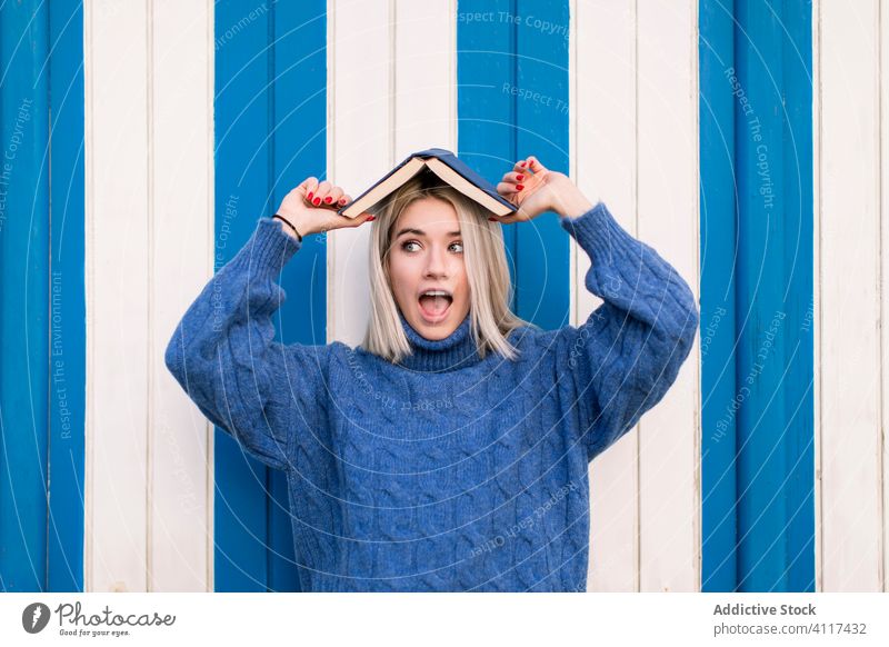 Amazed young woman with book on head fun amazed blue wall casual sweater knitted wow astonish color bright colorful female style trendy modern excited positive