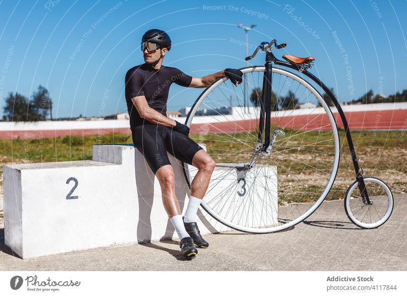 Pensive adult sportsman sitting alone on winner podium and leaning on penny farthing bike at sports stadium aspiration achievement high wheel bicycle challenge