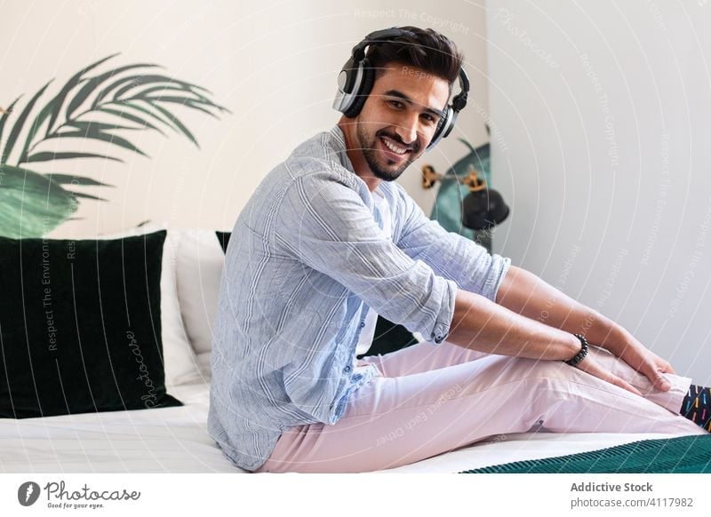 Ethnic man listening to music on bed home headphones ethnic cozy enjoy weekend male sit comfort melody tune audio song modern contemporary hispanic guy