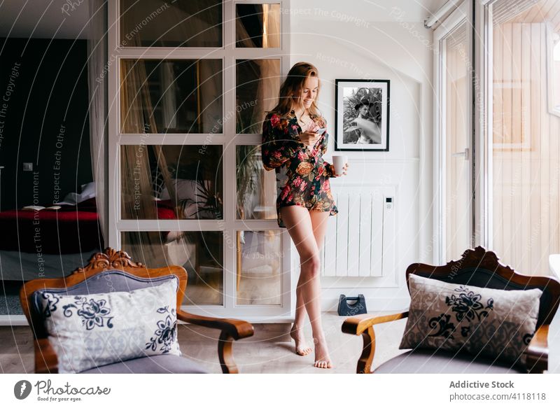 Woman drinking coffee and using smartphone at home woman morning enjoy window lingerie interior relax attractive female living room house pretty slim cup