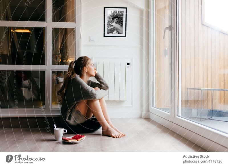 Dreamy woman having breakfast near window at home dream pensive floor toast coffee casual relax morning alone sit young female lifestyle rest serene tranquil