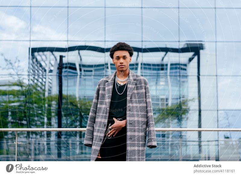 Stylish ethnic man in coat against glass wall trendy fashion urban street style chain accessory model city building serious modern african american black
