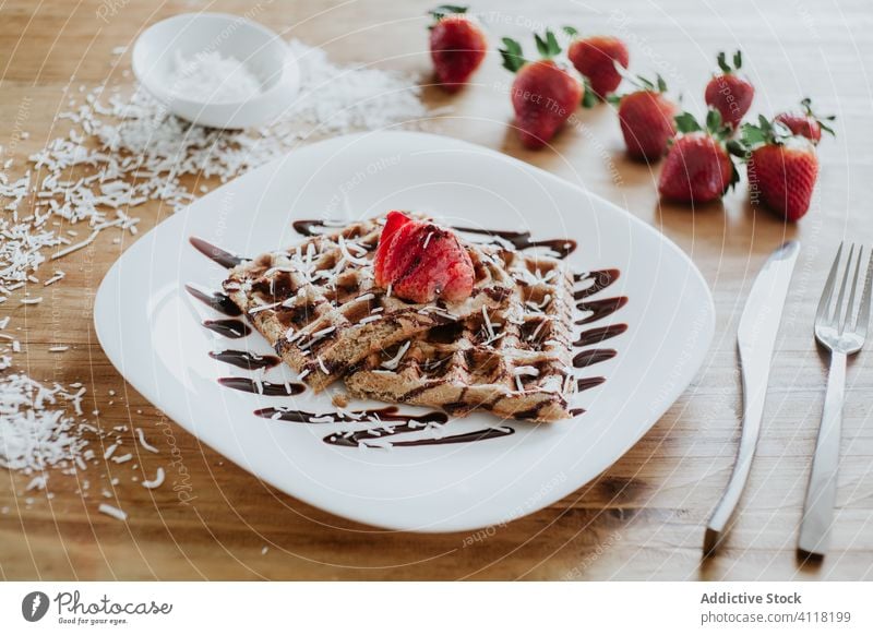 Waffles and strawberry with chocolate topping and coconut flakes waffle breakfast plate silverware table soft sweet yummy sauce wooden dessert delicious tasty