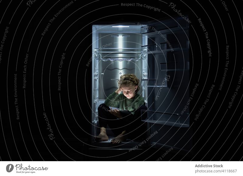 Calm relaxed kid sitting in empty fridge and using digital tablet at hot night eat child concept home refrigerator refreshment heat use chill device lounge