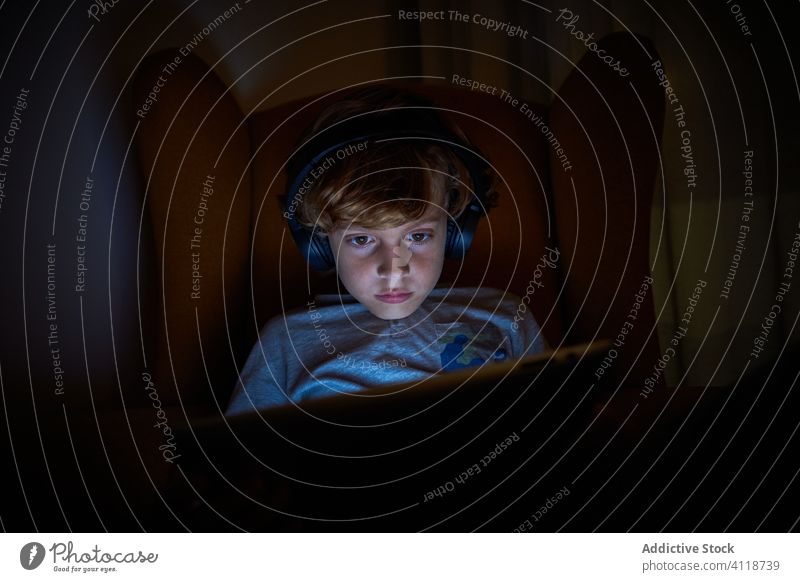 Child sitting with headphones on a sofa looking at the tablet at night boy interact facing device electronic imagination multimedia communication listen play