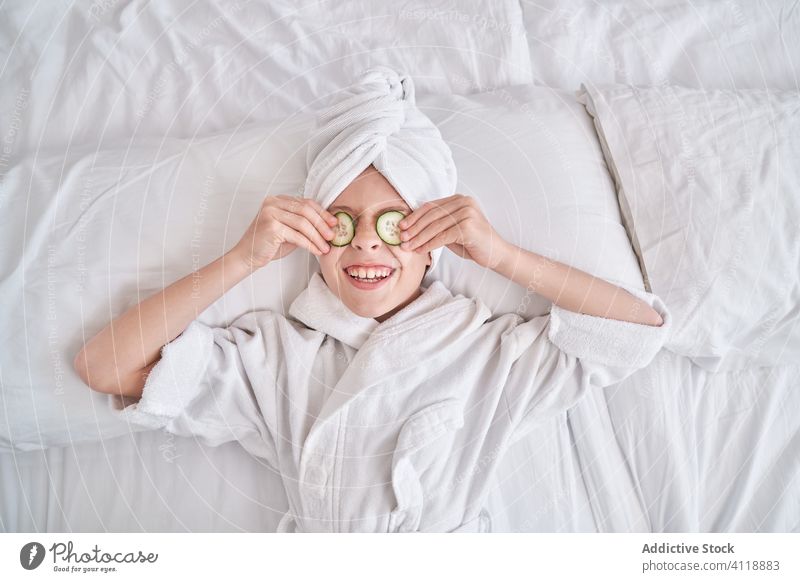 Cheerful kid with slices of cucumber on yes lying on bed mask beauty skin care fun positive laugh enjoy spa facial joke humor playful natural child happy