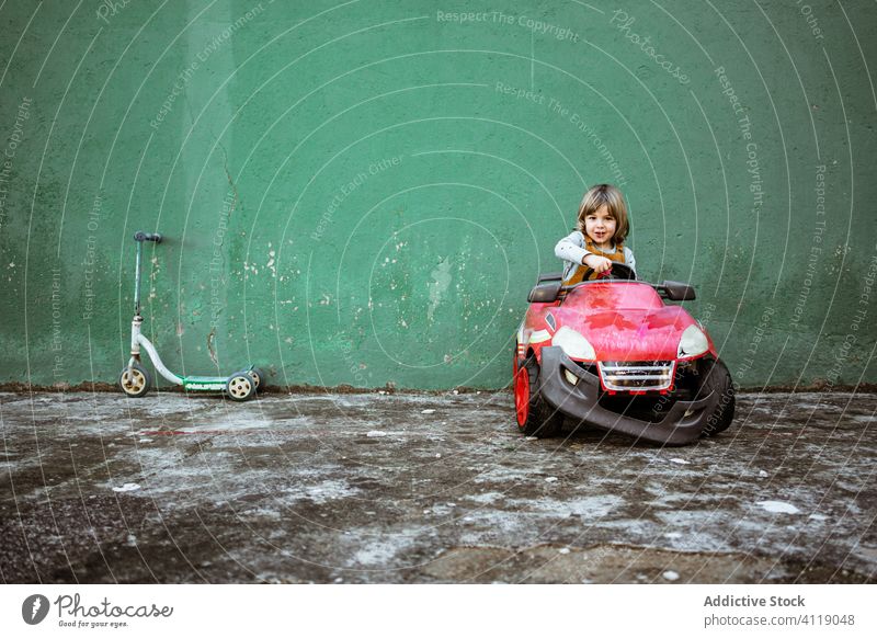 Little girl driving electric car drive toy play shabby wall broken bumper casual cute little child kid weathered damage rough grunge automobile childhood ride