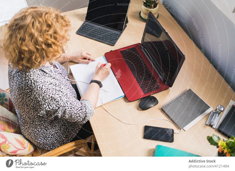 Anonymous woman taking notes in notebook while working on laptop smartphone workplace home freelance remote distance earphones notepad take note inspiration
