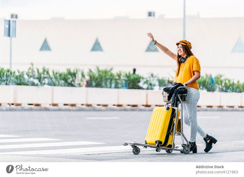 Happy woman with luggage cart catching taxi near airport hail road style building headphones transport passenger cap vehicle arrive trip city fashion yellow