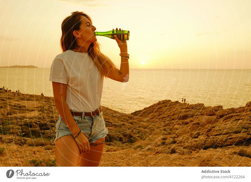 Smiling young lady with bottle of beer during sunset on seashore woman traveler smile refreshment summer chill leisure happy ibiza spain sunglasses drink
