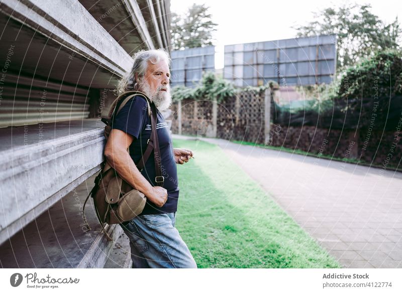 Aged traveling man smoking on the street traveler aged city male tourist elderly long hair gray hair senior casual wear backpack stand lean stone building