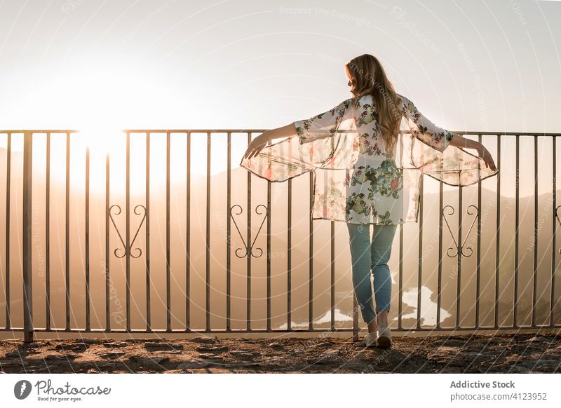 Anonymous woman standing near metal fence and enjoying picturesque view during sunset tourist sundown viewpoint admire terrace relax vacation evening freedom